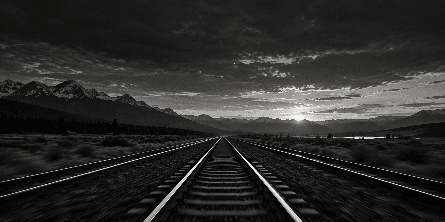 Black-and-white photograph of railway tracks running toward a low sun over distant mountains