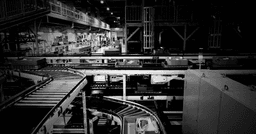 Dark, cinematic black-and-white photograph of an industrial warehouse conveyor-belt system moving packages